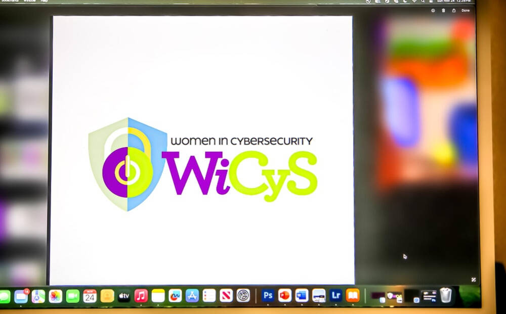 A computer screen displays the "Women in CyberSecurity (WiCyS)" logo, featuring a shield graphic and the text "WiCyS" in purple and lime green letters. The screen shows a Mac desktop with various app icons along the bottom.